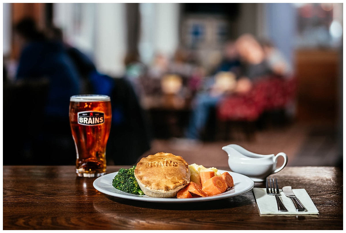 A photo of a pie with vegetables on a table with a pint of brains beer with a pub in the background